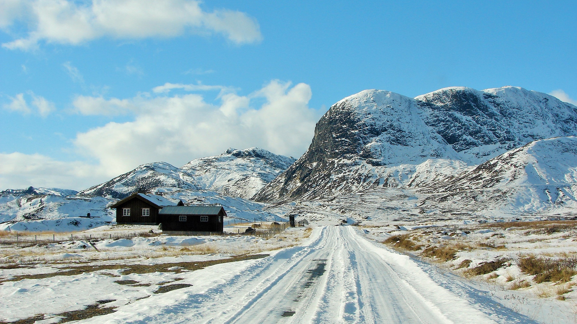 The snow covered road of Jotunheimvegen in wintertime passing mountains and two remote houses.