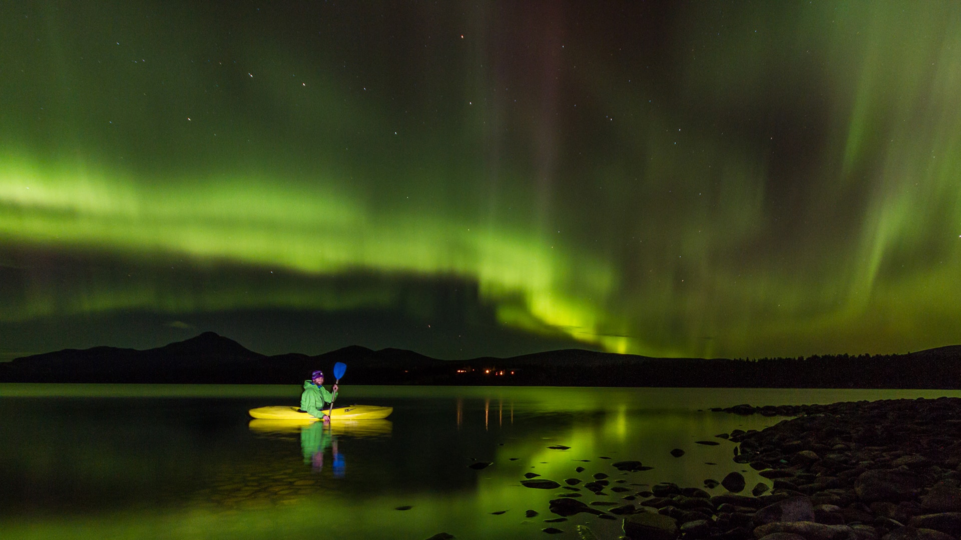 A person paddling in a kayak surrounded by northern lights reflecting in the lake.