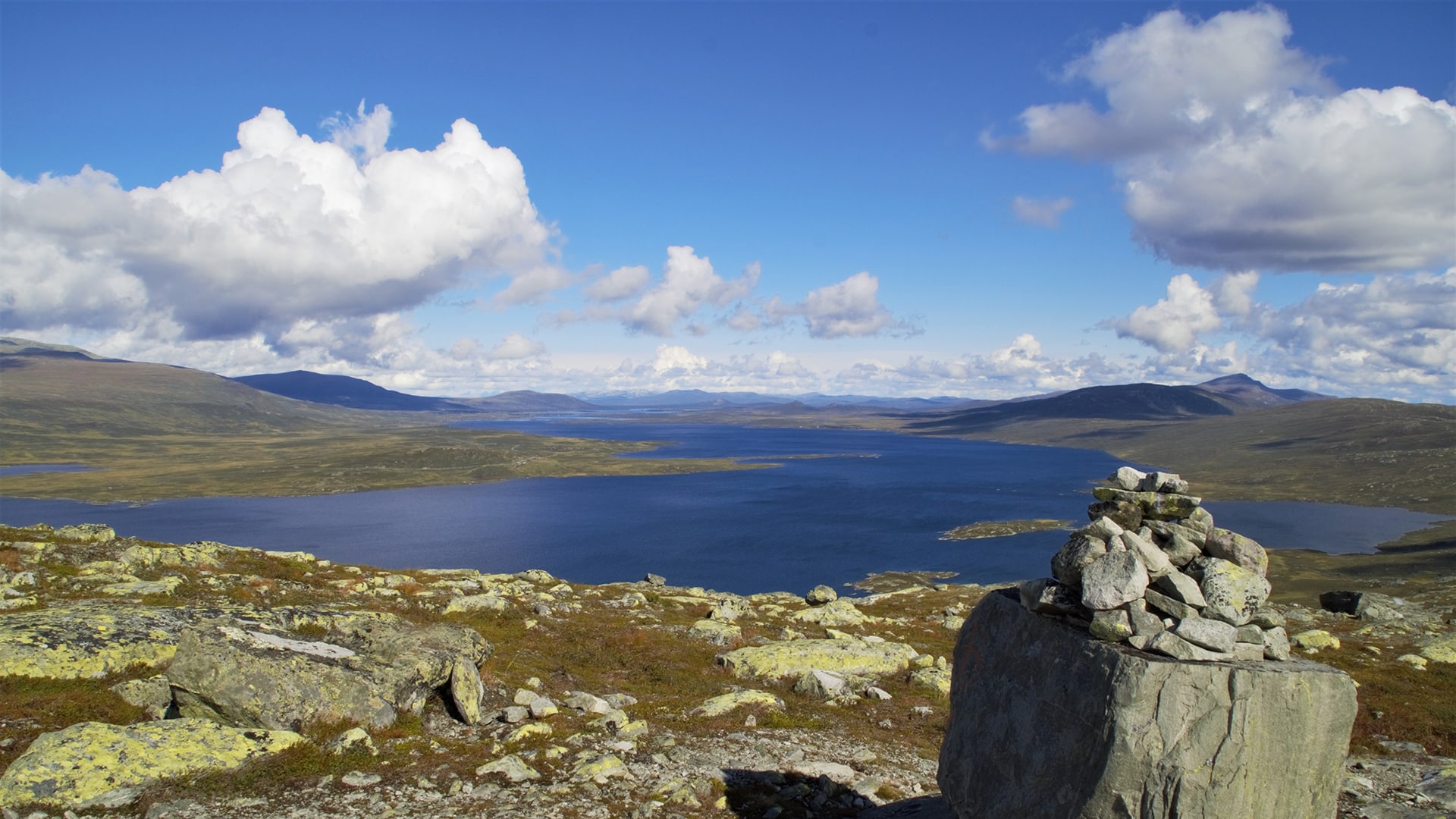 A stone cairn in front of Vinstervatn with tall mountains in the distance on a sunny summer day.