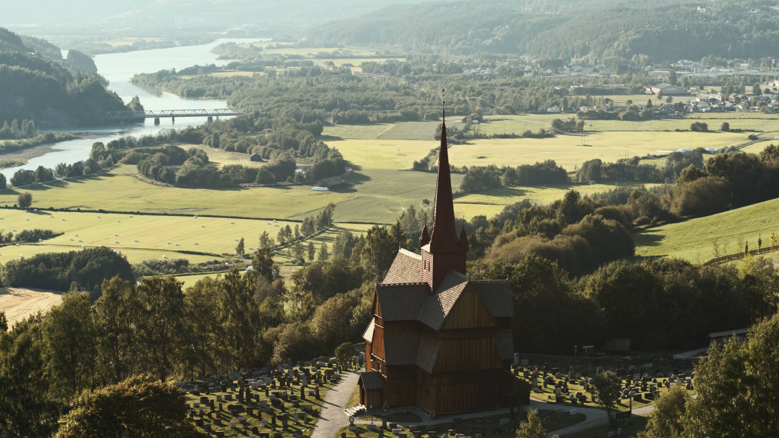 The stave church of Ringebu surrounded by fields and trees and a river flowing.